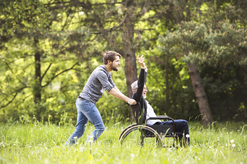 son walking with disabled father in wheelchair at park.