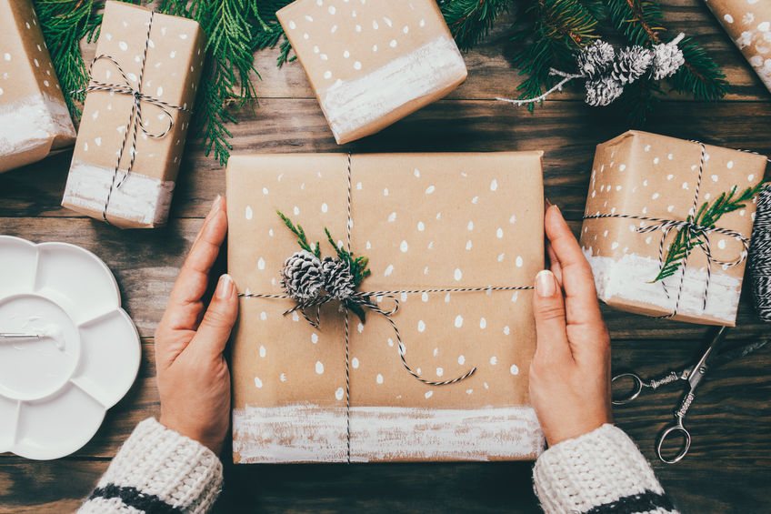 Woman´s hands wrapping Christmas presents on brown paper decorated with painted snow, fir branches and pinecones on a rustic wooden board. Top view