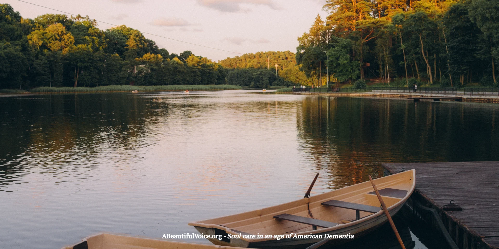 Peaceful Midwestern Lake at dusk, similar to the lake area where I took my parents for picnics in the early stage of their dementia. The sun is shining on the upper parts of the trees surrounding the lake.